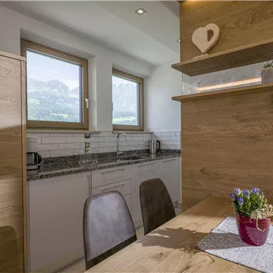 A modern kitchen with wooden details and a dining area. Large windows provide a view of the mountains.
