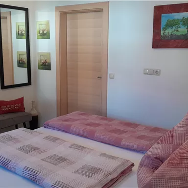 A cozy bedroom with two beds and red pillows. On the wall hang pictures and a mirror, there is a small television and a wooden door.