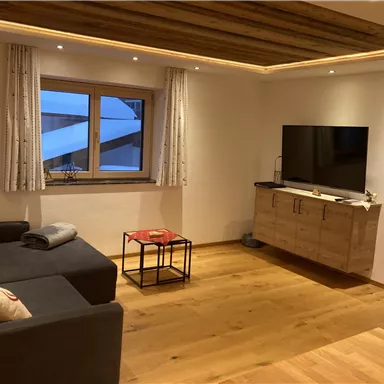 A cozy living room with a gray sofa and a TV on the wall. The room features wooden details and large windows that let in daylight.