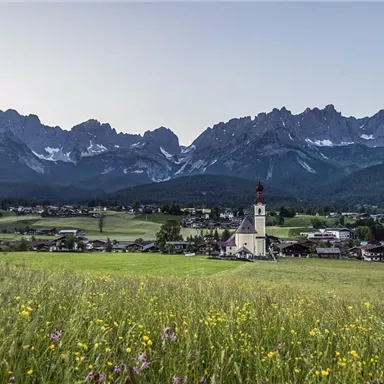 A picturesque village with a church in the foreground and impressive mountains in the background. The meadow is covered with colorful flowers and radiates a peaceful atmosphere.