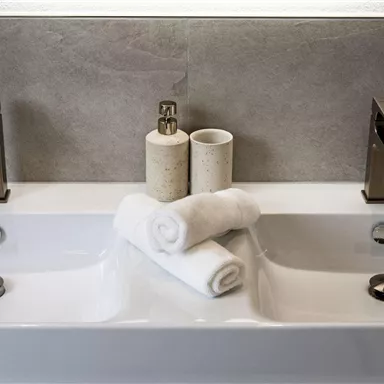 Ein modernes Badezimmer mit zwei Waschbecken und eleganten Wasserhähnen. Saubere, gefaltete Handtücher liegen in der Mitte und sorgen für eine entspannte Atmosphäre.