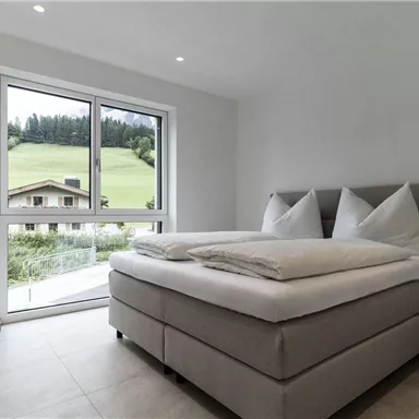 Ein modernes Schlafzimmer mit einem großen Fenster, das einen Blick auf grüne Hügel bietet. Das Zimmer ist hell und freundlich mit einem bequemen Bett und minimalistischer Einrichtung.