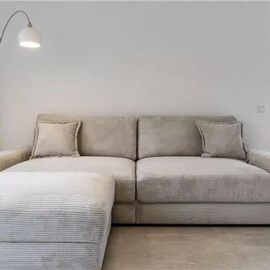 Ein modernes, helles Wohnzimmer mit einem beigen Sofa und einer eleganten Stehlampe. Im Hintergrund sind Fenster mit Blick auf eine grüne Landschaft und Berge.