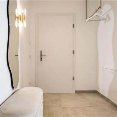 Ein heller Flur mit einem runden Spiegel und einer weißen Bank. An der Wand hängt ein Kleiderbügel und die Tür ist geschlossen.
