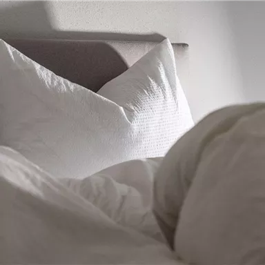 Ein gemütliches Schlafzimmer mit weißen Kissen und einer weichen Decke. Sanftes Licht fällt auf das Bett.