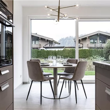 Eine moderne Küche mit einem runden Tisch und gepolsterten Stühlen. Der Blick geht auf ein grünes Grundstück und bergige Architektur im Hintergrund.