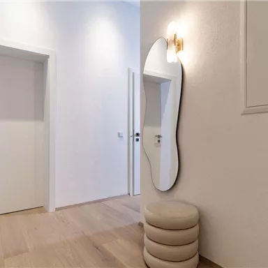 Ein heller Flur mit modernen, weißen Türen und einem eleganten Spiegel. Der Boden ist aus hellem Holz und es gibt eine stilvolle Sitzgelegenheit.