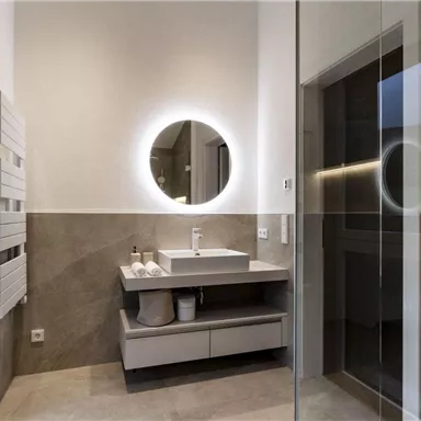 Ein modernes Badezimmer mit einem schlichten Waschbecken und einem dazu passenden runden Spiegel. Die Wände sind in neutralen Tönen gehalten und das Licht ist hell und einladend.
