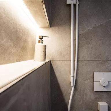 Ein modernes Badezimmer mit grauen Fliesen und sanfter Beleuchtung. Auf einer Ablage steht ein eleganter Seifenspender.