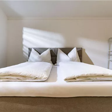 Ein modernes Schlafzimmer mit einem bequemen Doppelbett und frischen weißen Bettdecken. Sanftes Licht fällt durch die Fenster und schafft eine gemütliche Atmosphäre.