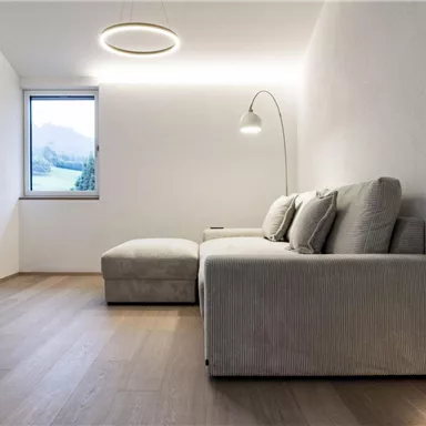 Ein modernes Wohnzimmer mit einem beigen Sofa und einem großen Fenster. An der Wand hängt ein Fernseher, und es gibt eine stehlampe für zusätzliche Beleuchtung.