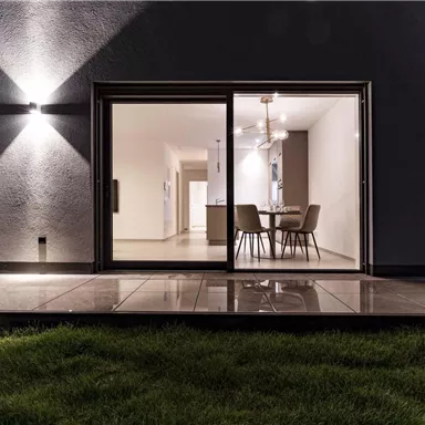 Ein modernes Wohnzimmer mit großen Fenstern, das auf eine Terrasse mit Grasblick führt. Die Beleuchtung schafft eine einladende Atmosphäre in der Abendstimmung.