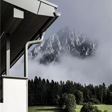 Ein Blick auf die Berge mit Nebel und grünen Wiesen im Vordergrund. Die Architektur des Daches fügt sich harmonisch in die Landschaft ein.