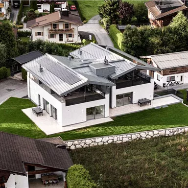 Ein modernes Haus mit einem flachen Dach und großen Fenstern. Die Umgebung ist grün und gepflegt, mit einigen anderen Gebäuden in der Nähe.