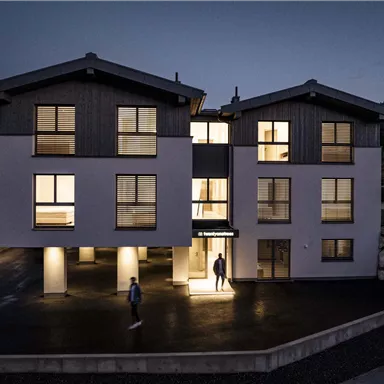 Ein modernes Gebäude bei Nacht, beleuchtet mit warmem Licht. Menschen bewegen sich vor dem Eingang und die Umgebung ist ruhig.