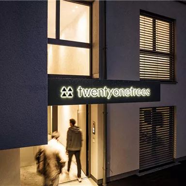 Ein modernes Gebäude mit dem Eingang von "twentyone trees". Zwei Personen betreten das Gebäude bei Abendbeleuchtung.