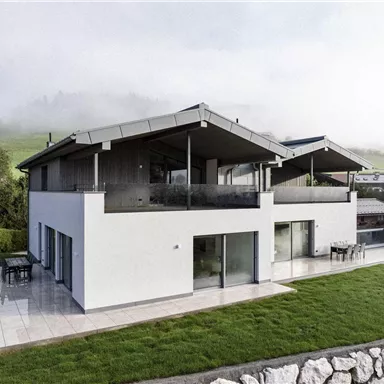 Ein modernes Haus mit großen Fenstern und einem Terrassenbereich. Die grüne Rasenfläche und die umgebende Landschaft schaffen eine ruhige Atmosphäre.