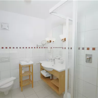A modern bathroom with white tiles and a clear shower area. There is a wooden shelf with towels and a stylish sink setup.