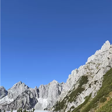 Eine beeindruckende Berglandschaft mit steilen Felsen und grünem Gelände. Der Himmel ist klar und blau, während Wolken im Tal liegen.