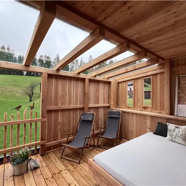 A cozy wooden room with a bed and two loungers. The view overlooks a green meadow and trees.