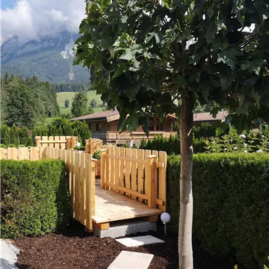 Eine stilvolle Holzbrücke führt durch eine grüne Heckenlandschaft. Im Hintergrund sind Berge und ein blauer Himmel zu sehen.