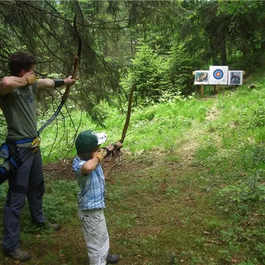 An adult and a child practice archery in the forest. In the background, target boards can be seen.