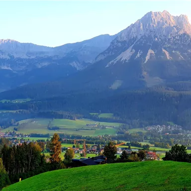 Eine wunderschöne Berglandschaft mit grünen Wiesen und bewaldeten Hügeln. Im Hintergrund erheben sich majestätische Berge unter einem klaren Himmel.