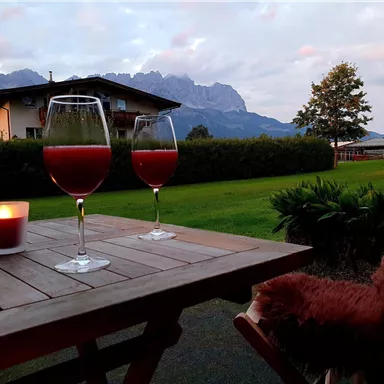 Ein Tisch mit zwei Gläsern rotem Getränk und einer Kerze steht vor einer malerischen Berglandschaft. Im Hintergrund sind Bäume und ein Gebäude zu sehen, unter einem bewölkten Himmel.