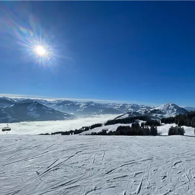 A snow-covered mountain landscape under a clear blue sky. The sun shines over the peaks and the clouds lie low in the valley.
