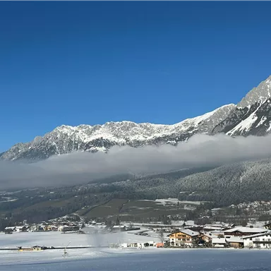 A snowy winter landscape with mountains in the background. In the foreground lies a small village, surrounded by a white blanket of snow.
