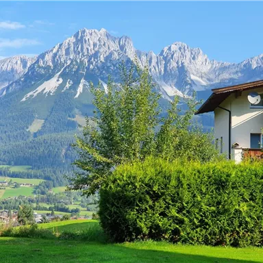 Ein malerischer Bergblick mit majestätischen Gipfeln und grünem Gras im Vordergrund. Im Bild ist ein ruhiges Wohngebiet mit Bäumen und einer klaren blauen Himmelsfarbe.