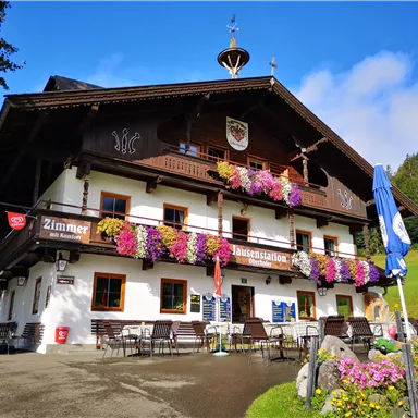 Ein traditionelles Alpenhaus mit bunten Blumen am Balkon. Es gibt Sitzgelegenheiten im Freien und eine klare Berglandschaft im Hintergrund.