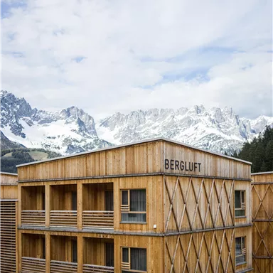A modern wooden building with the inscription "BERGLUFT" in front of majestic mountains. The sky is slightly cloudy and the landscape appears calm and inviting.