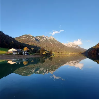 Ein ruhiger See umgeben von Bergen und Wäldern. Der Himmel ist klar und spiegelt sich im Wasser.