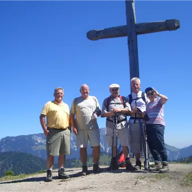 A group of five hikers stands in front of a large wooden cross in the mountains. The surroundings display an impressive landscape with mountains and a clear sky.