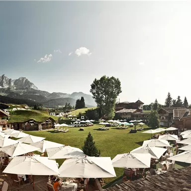 A beautiful hotel with a large garden and umbrellas. In the background, mountains and a clear sky can be seen.