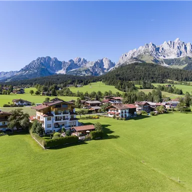 A picturesque landscape with green meadows and charming buildings. In the background, impressive mountains rise under a clear sky.