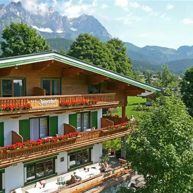 A charming wooden house with flower boxes and many windows. In the background, majestic mountains rise under a clear sky.