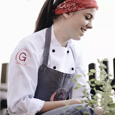 A cook with a red bandana is kneeling and picking herbs. She is wearing a white chef's jacket and is surrounded by plants.