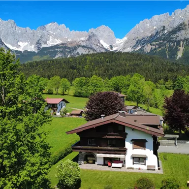 A picturesque landscape with a charming alpine chalet and green grass. In the background, majestic mountains rise under a clear blue sky.