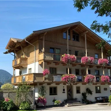 A charming wooden house in alpine style with balconies adorned with colorful flowers. In the background, gentle mountains and a clear blue sky can be seen.