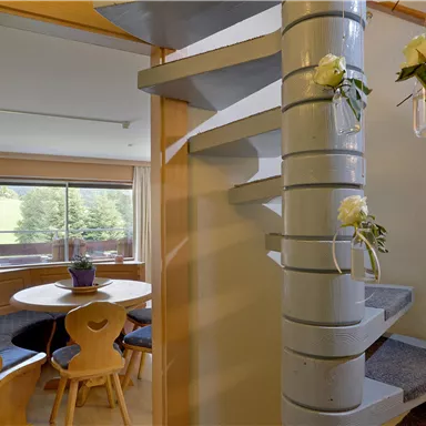 Ein heller Eingangsbereich mit einer geschwungenen Treppe und Blumen in Glasvorrichtungen. Im Hintergrund ist ein gemütlicher Essbereich mit einem Fenster und Blick ins Freie zu sehen.