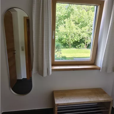 A bright room with a window overlooking a green meadow. Next to the window stands a simple wooden bench and a rectangular mirror.