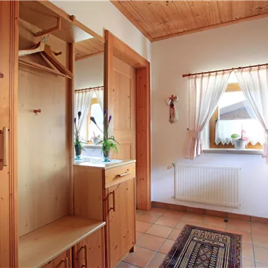 Ein heller Flur mit Holzschränken und einem Fenster mit Vorhängen. Der Boden ist gefliest und es liegt ein Teppich aus.