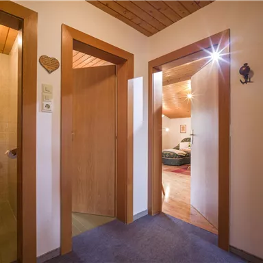 Ein flacher Flur mit hölzernen Türen und einem Teppichboden. Im Hintergrund ist ein gemütliches Zimmer zu sehen.