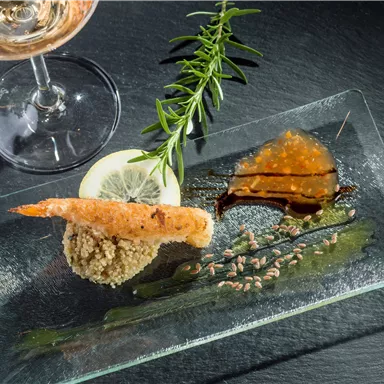 An artfully arranged plate with a crab, lemon slice, and sauces. In the background, there is a glass of rosé wine and a sprig of rosemary.