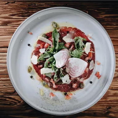 A delicious dish on a white plate, consisting of raw meat, salad, and various toppings. The background is made of light wood.