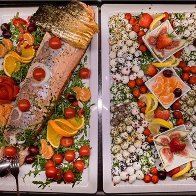 Ein reichhaltiges Buffet mit gegrilltem Lachs, frischen Beilagen wie Tomaten und Zitrusfrüchten. Auf der anderen Seite sind ansprechend dekorierte Platten mit Obst und leckeren kleinen Häppchen zu sehen.