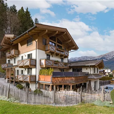 A charming wooden house in the mountains with several balconies. The surroundings are filled with green meadows and trees.