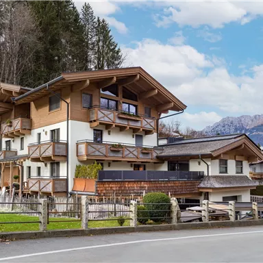 A modern alpine house with several balconies and a wooden facade. In the background, gentle mountains and a clear sky can be seen.
